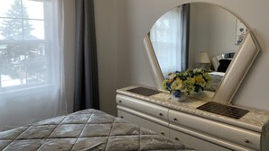 Ett sovrum med en säng, en byrå med en spegel, ett fönster med gardiner och en vas med blommor på byrån.