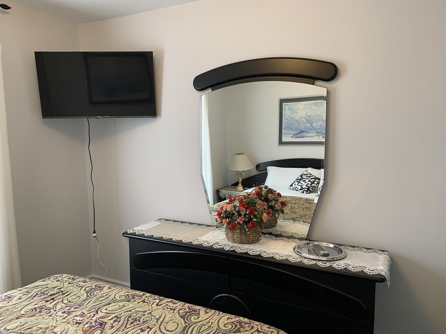 A bedroom with a bed, a dresser, a mirror, a TV mounted on the wall, and a bedside table with a lamp and a framed picture.