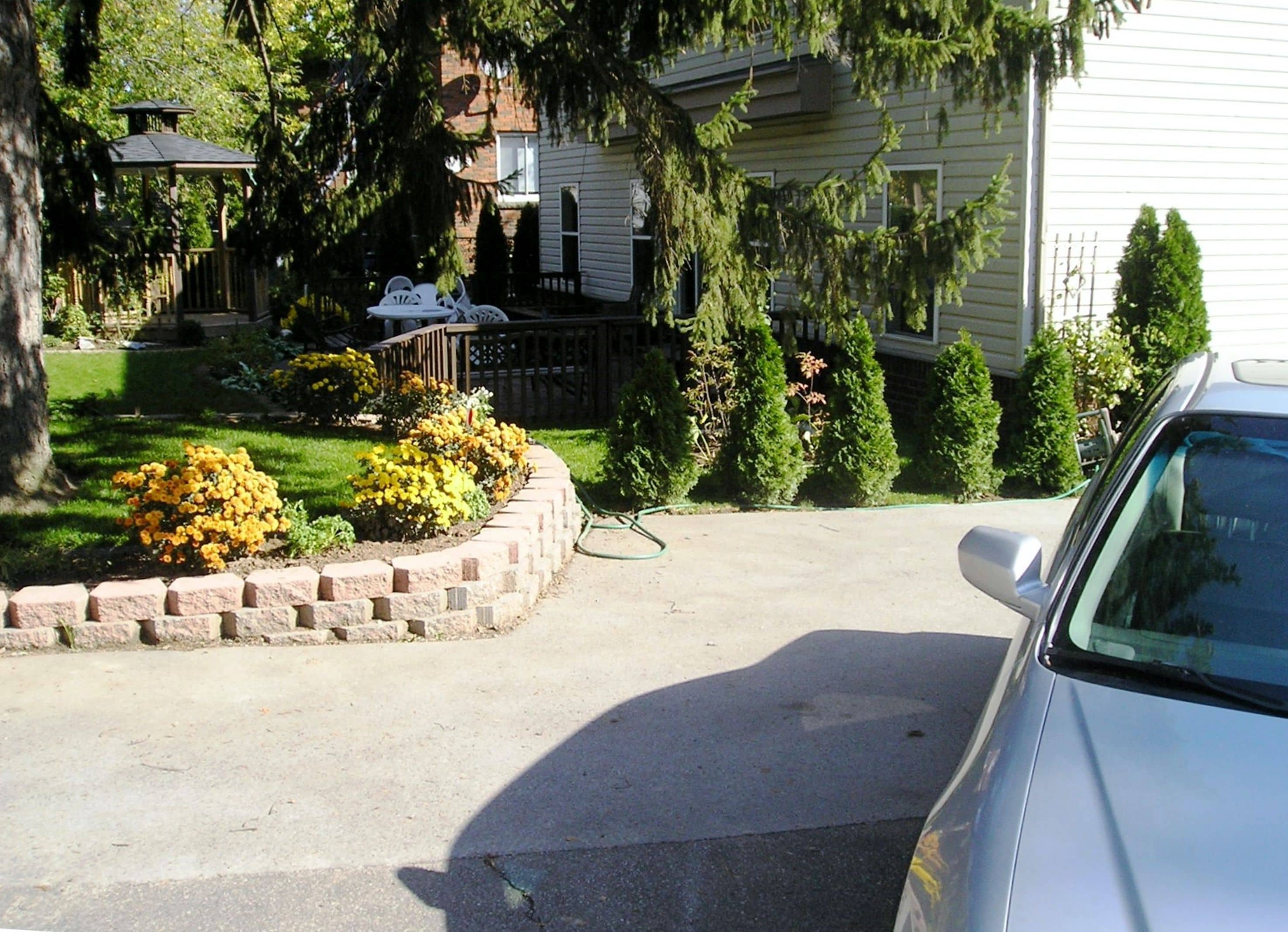 Un camino de acceso residencial con un automóvil estacionado a la derecha. Hay un jardín pequeño con flores amarillas y un borde de ladrillo. En el fondo se ve una terraza de madera y una silla de patio.