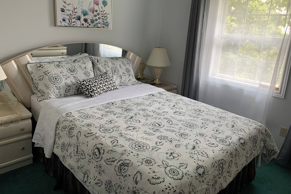 Una cama bien hecha con ropa de cama floral, una mesita de noche con lámpara y una ventana con cortinas.