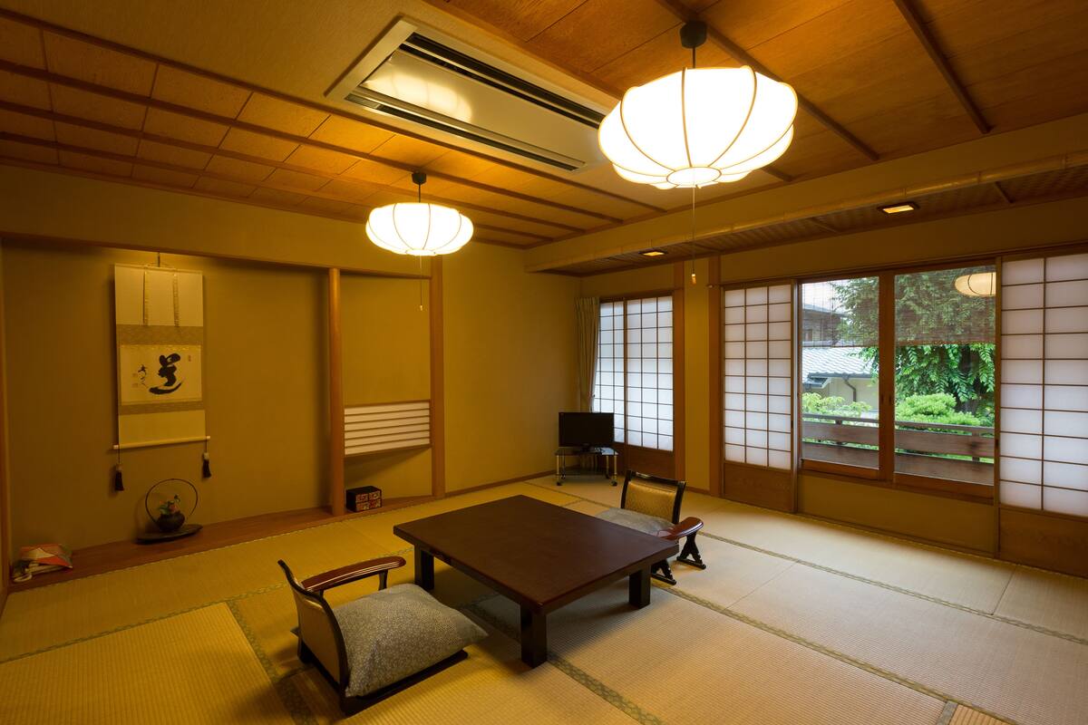 Um quarto japonês tradicional com piso de tatame, uma mesa baixa, um assento almofadado, uma televisão e vista para o verde lá fora.