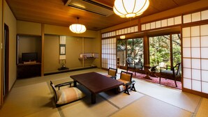A traditional Japanese room with tatami flooring, a low table, and chairs. There is a television on a stand, a sliding door leading to an outdoor area, and a large window with a view of greenery. - Arashiyama Benkei (Kyoto)