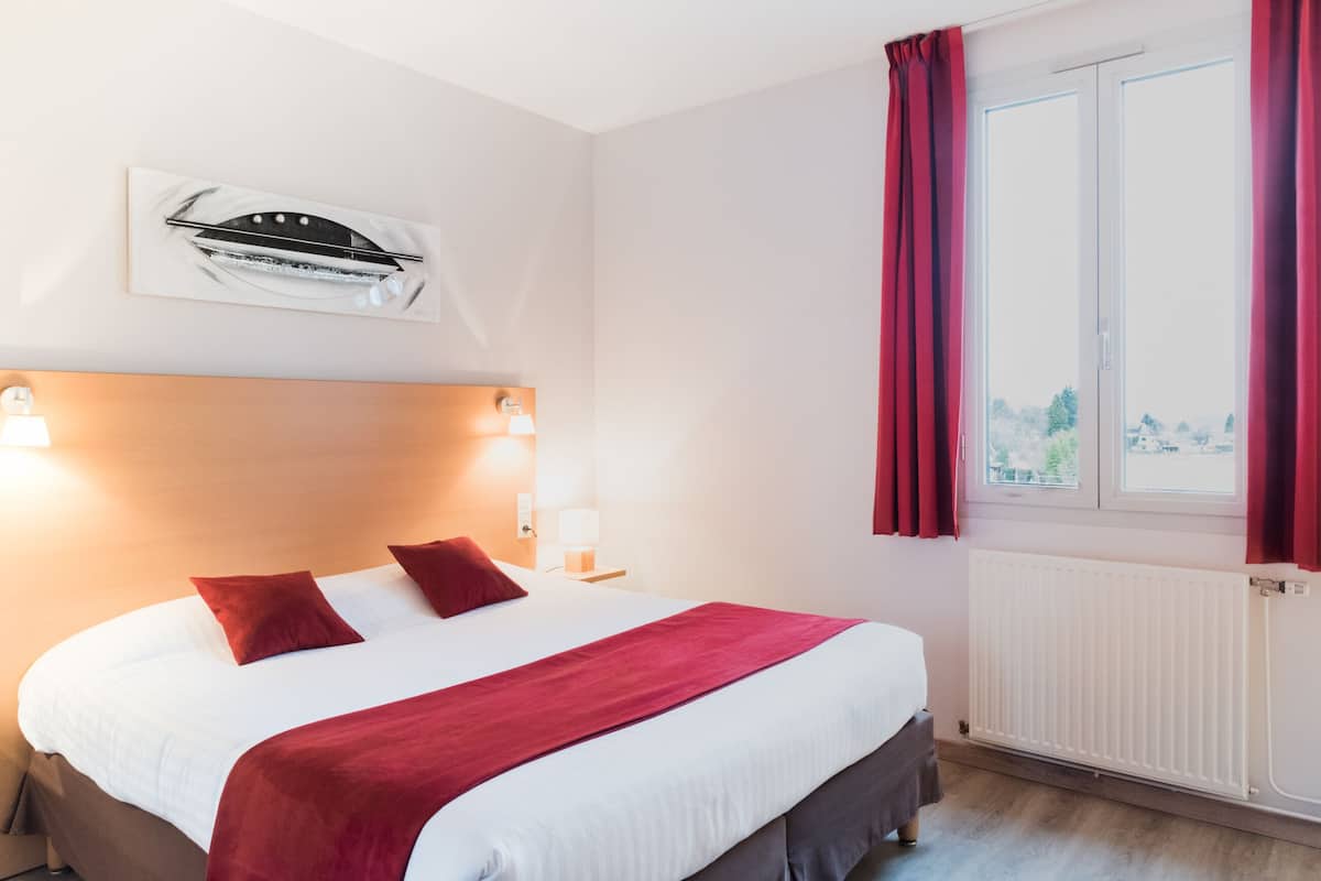 Quarto de hotel com uma cama grande, duas luminárias de cabeceira, uma janela com cortinas vermelhas e um radiador.