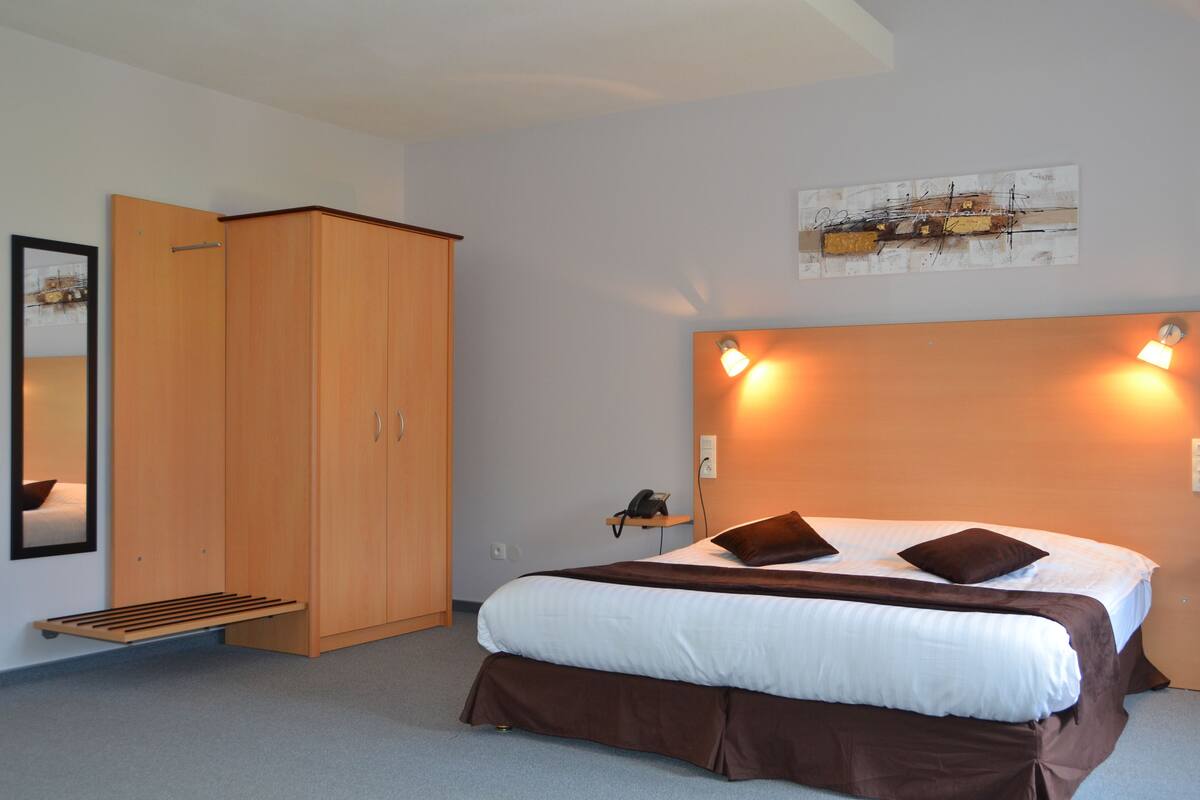 Quarto de hotel com cama, guarda-roupa, mesa de cabeceira com abajur e uma obra de arte na parede.