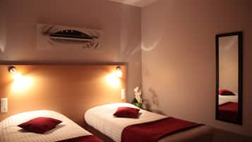 A hotel room with two beds, each with a red pillow and a white bedspread. There is a wall-mounted air conditioning unit above the beds, and a mirror on the wall to the right.