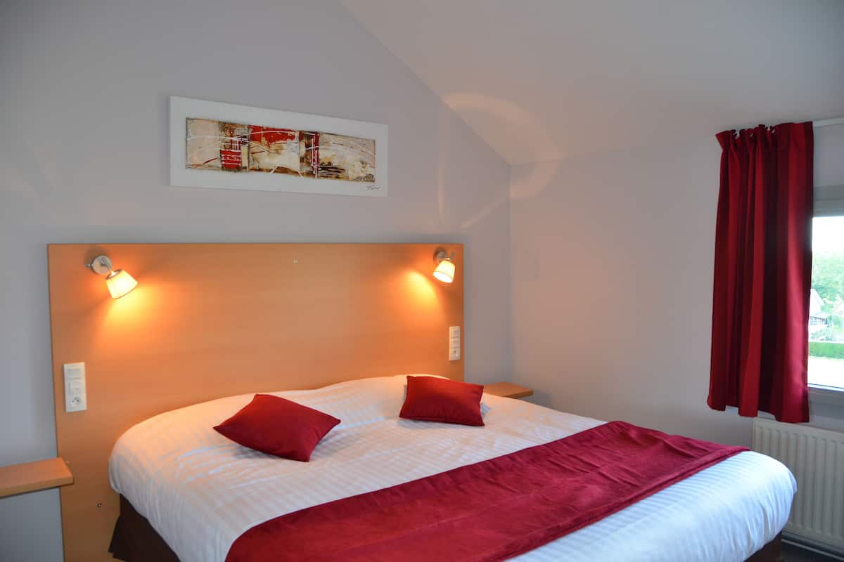 Quarto de hotel com uma cama grande, duas almofadas vermelhas, um cobertor vermelho, cabeceira com luminárias fixadas na parede, uma obra de arte emoldurada acima da cama e uma janela com cortinas vermelhas.