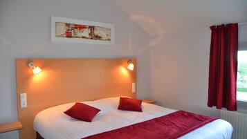 A hotel room with a large bed, two red pillows, a red throw blanket, a headboard with wall-mounted lights, a framed artwork above the bed, and a window with red curtains.