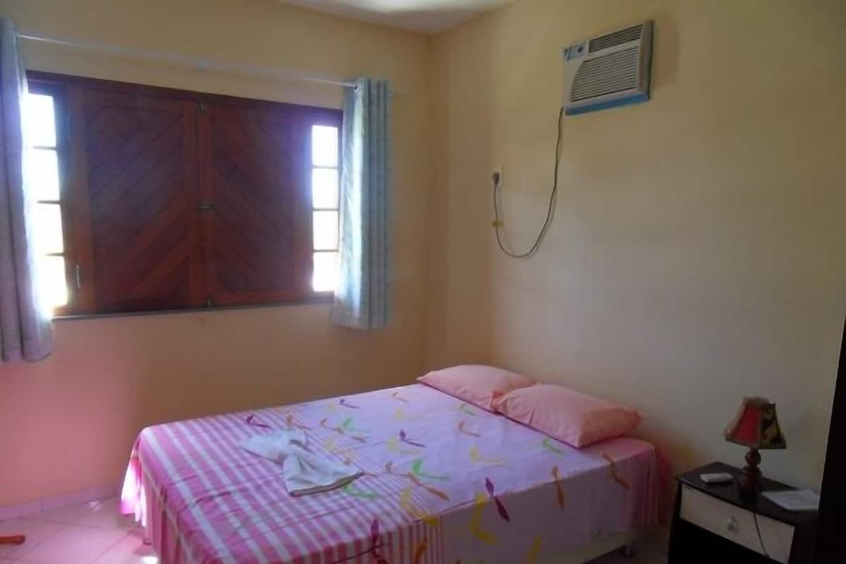 Een slaapkamer met een bed, nachtkastje, raam met gordijnen en een airconditioning.