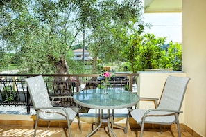 A balcony with a round glass table and two wicker chairs, surrounded by trees and a view of buildings in the background. - Lemon Garden Villa (Kassandra)