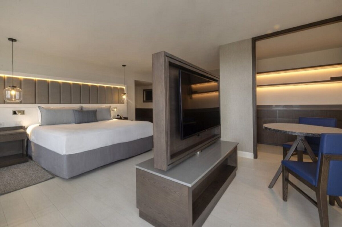 A modern bedroom with a large bed, a TV on a stand, and a dining area with a table and chairs.