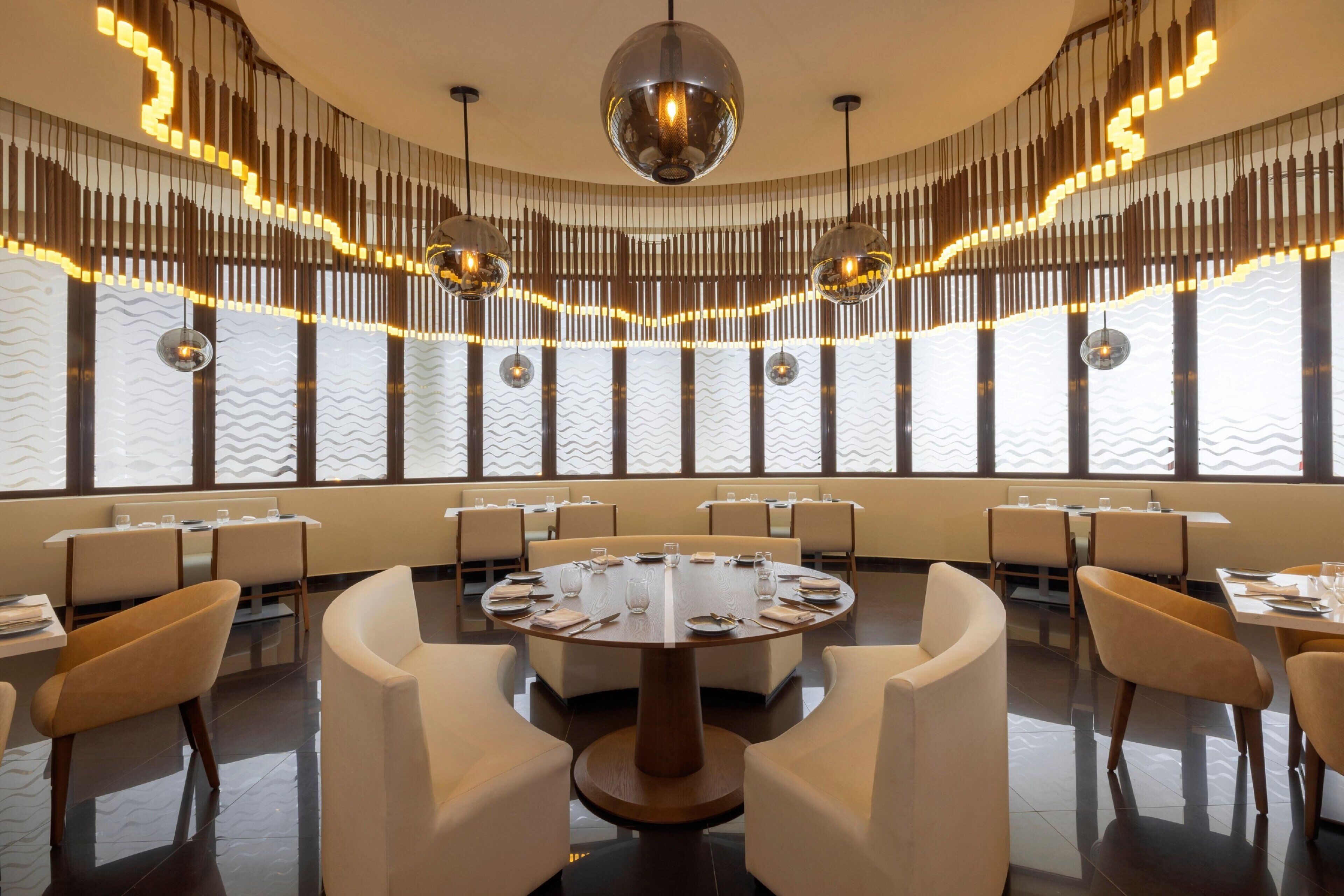 A modern dining area with round tables and high-backed chairs, large windows, and decorative lighting.