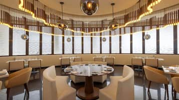 A modern dining area with round tables and high-backed chairs, large windows, and decorative lighting.