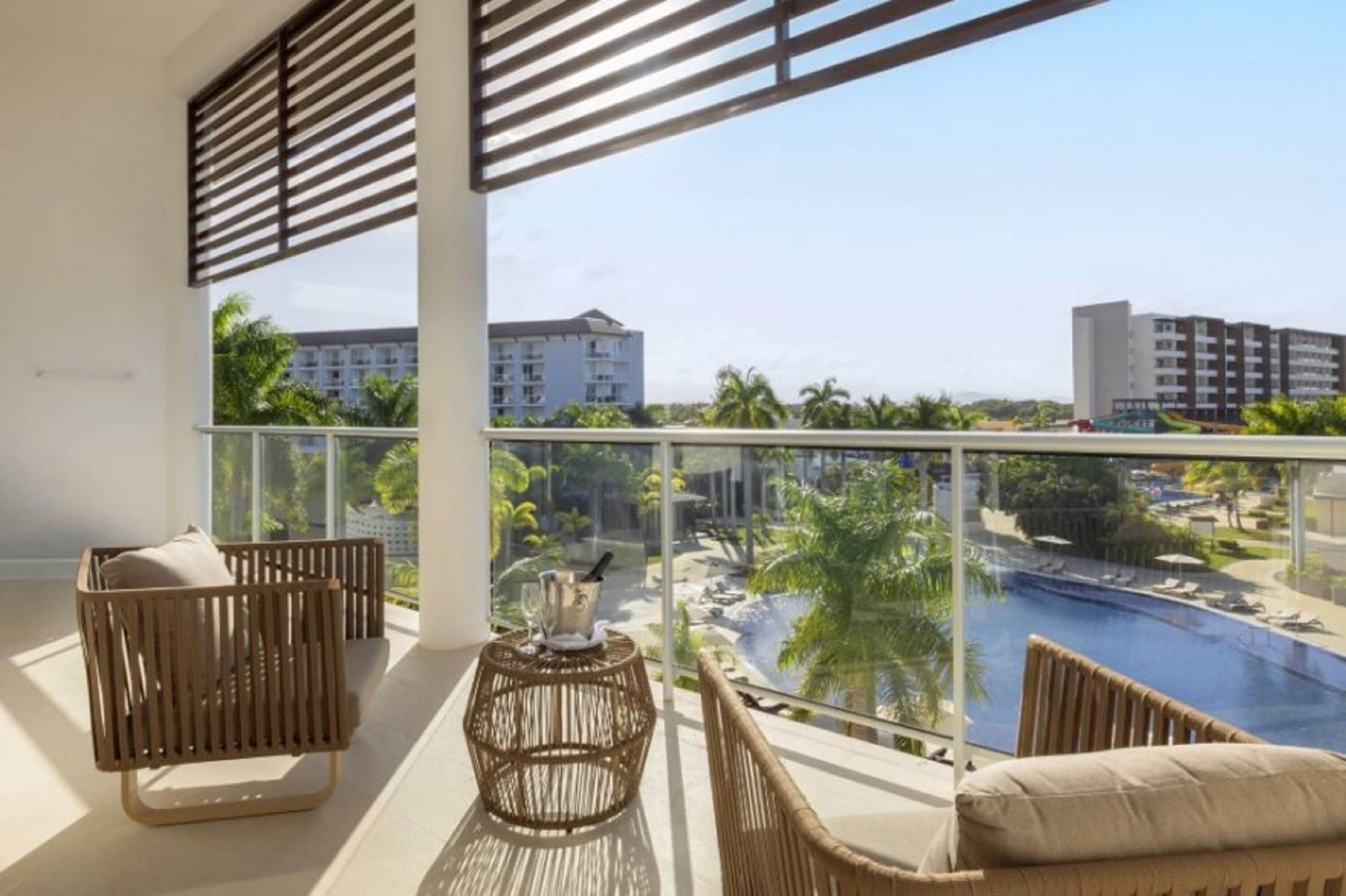 A balcony with wicker furniture, a glass table, and a view of a pool and buildings.