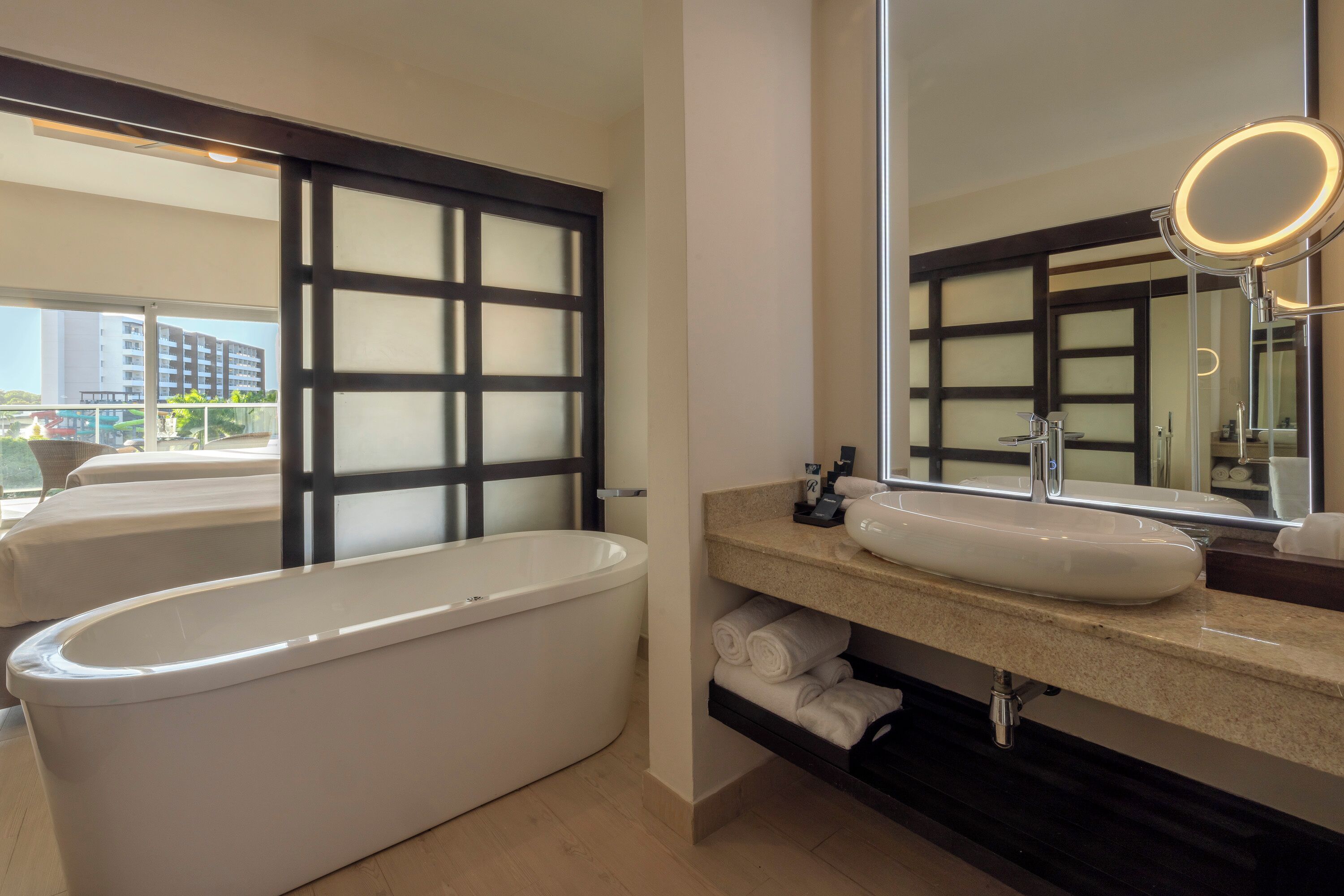 A modern bathroom with a freestanding bathtub, a large mirror with a round light, and a view of the cityscape through a large window.
