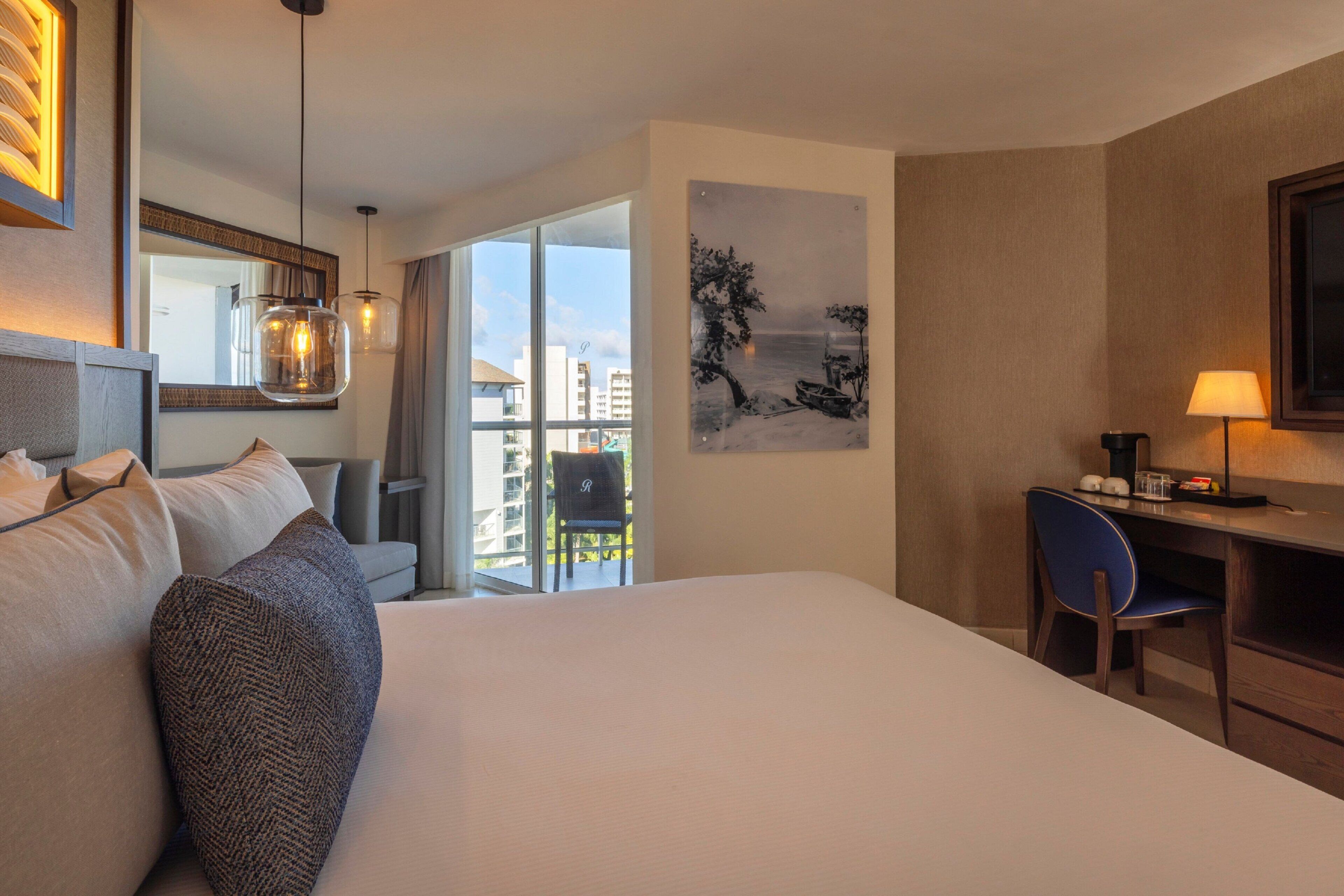 A hotel room with a large bed, a desk with a chair, a desk lamp, a painting on the wall, and a view of buildings through the window.