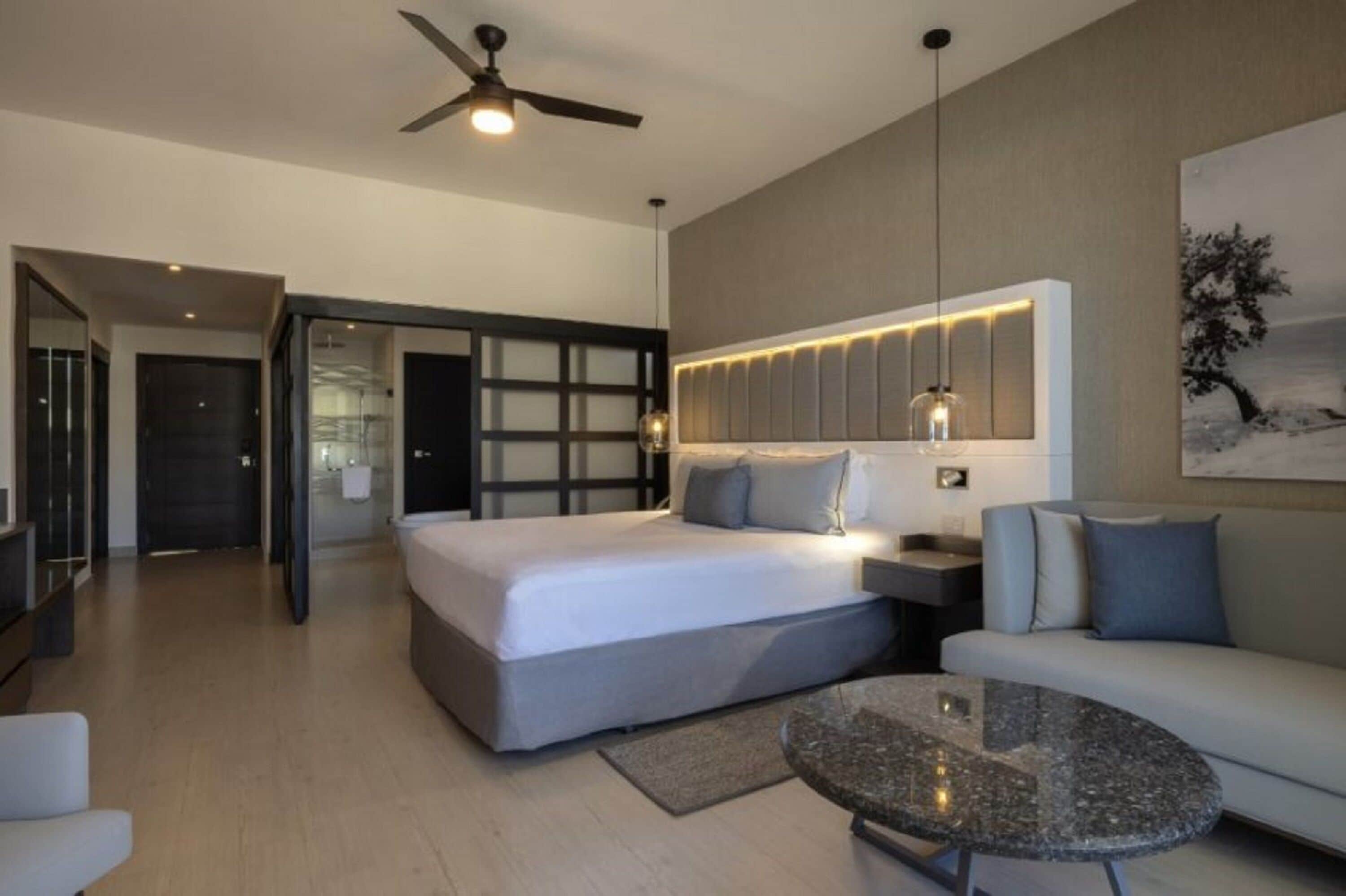 A modern hotel room with a large bed, a sofa, a round coffee table, and a ceiling fan.