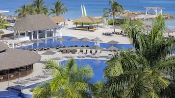 Un complejo turístico con una gran piscina, zonas de descanso y una playa con sombrillas y palmeras.