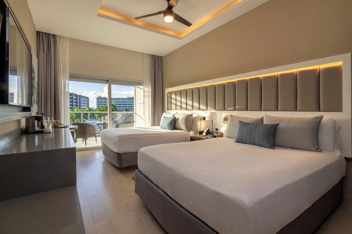 A modern hotel room with two beds, a desk, and a balcony with a view of buildings.