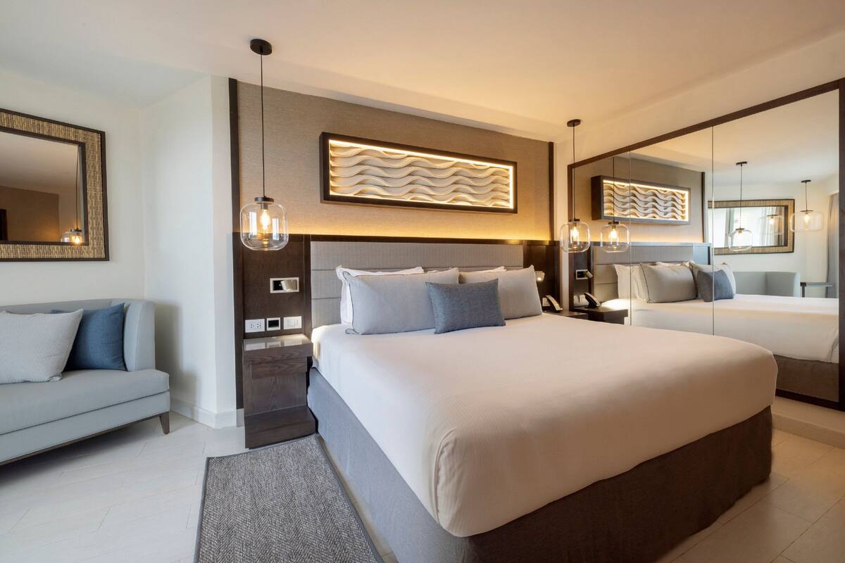 A modern hotel room with a large bed, a sofa, a bedside table, a mirror, and a wall-mounted light fixture.