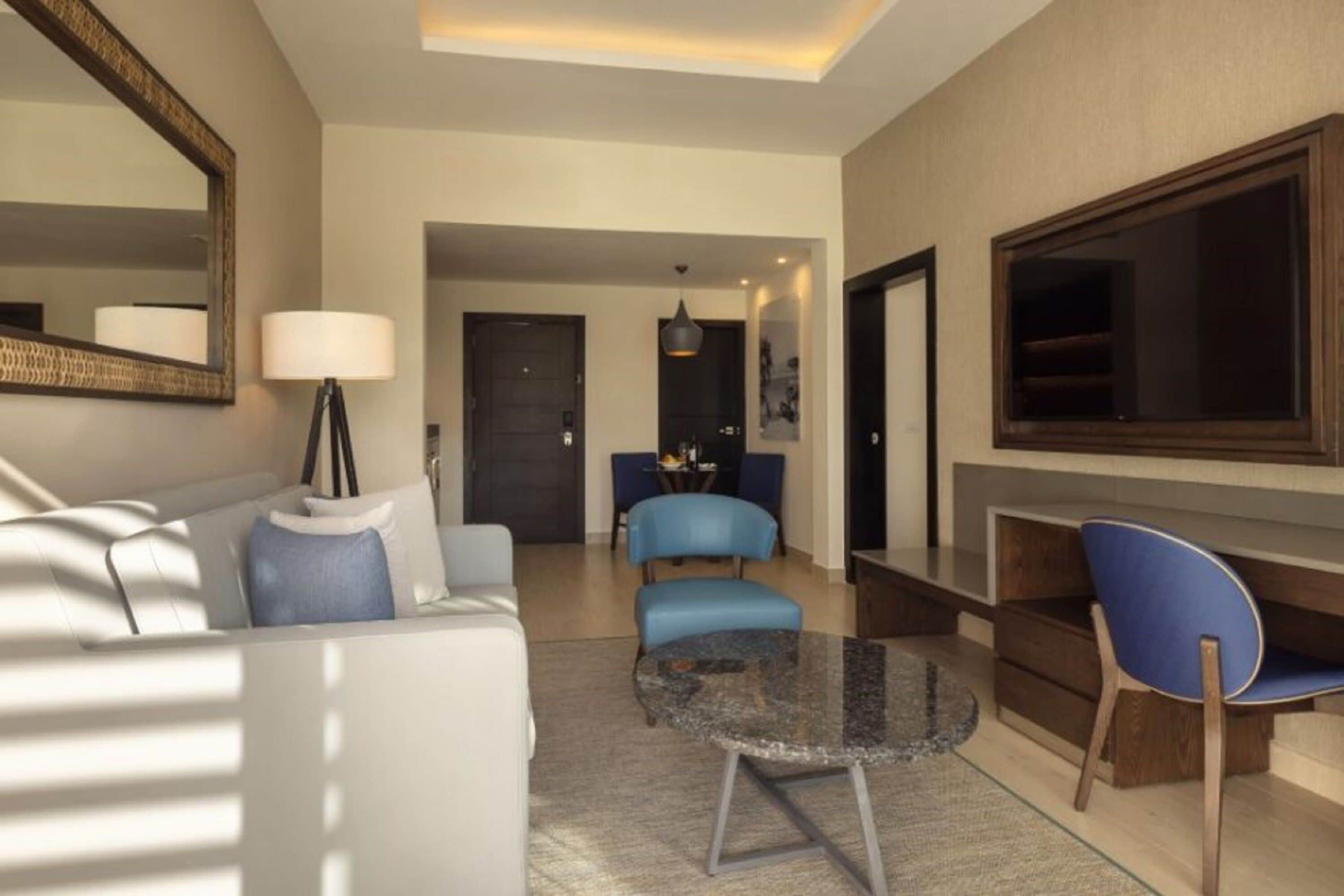 Een moderne hotelkamer met een bank, een ronde tafel en een televisie. Er is een deur die naar een andere kamer leidt en een raam met gordijnen.