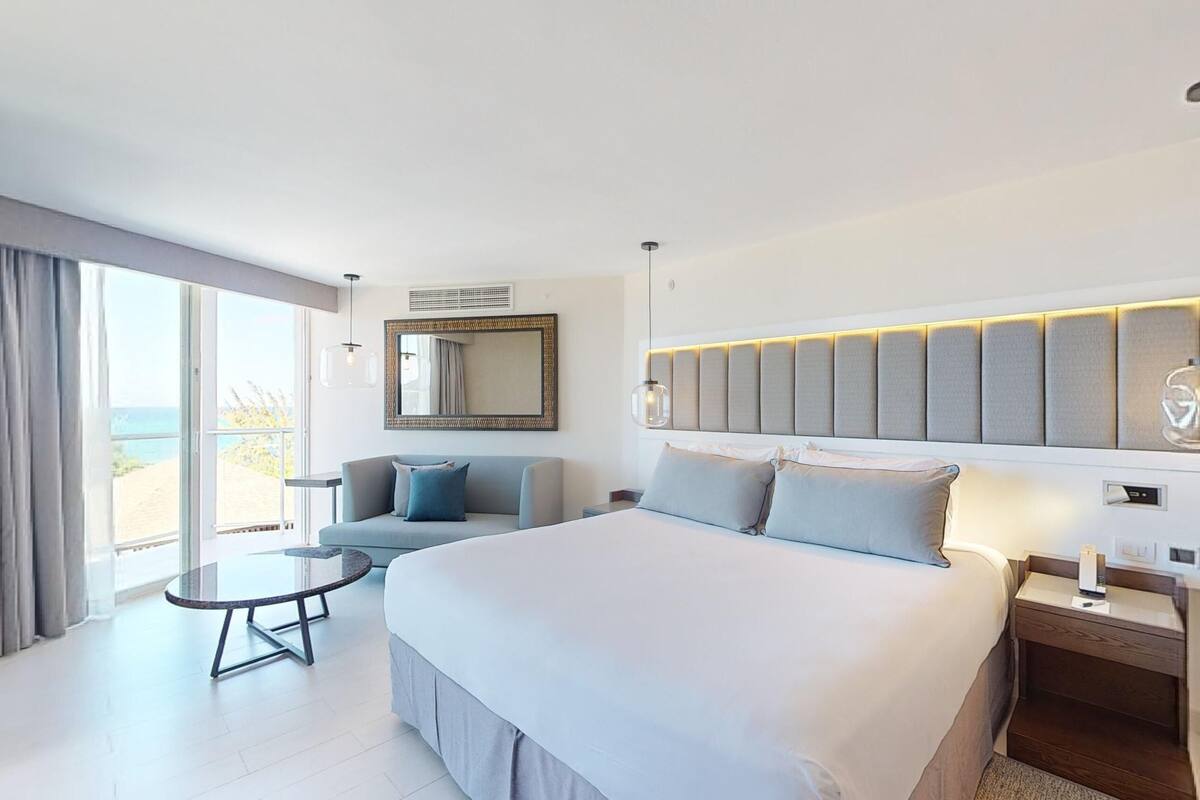 A modern hotel room with a large bed, a sofa, a small table, and a view of the sea.