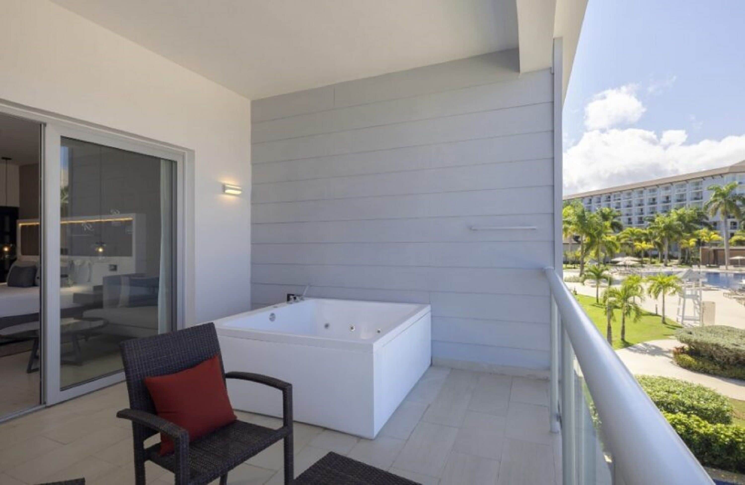 A balcony with a white bathtub, a wicker chair with a red cushion, and a view of a hotel pool and palm trees.