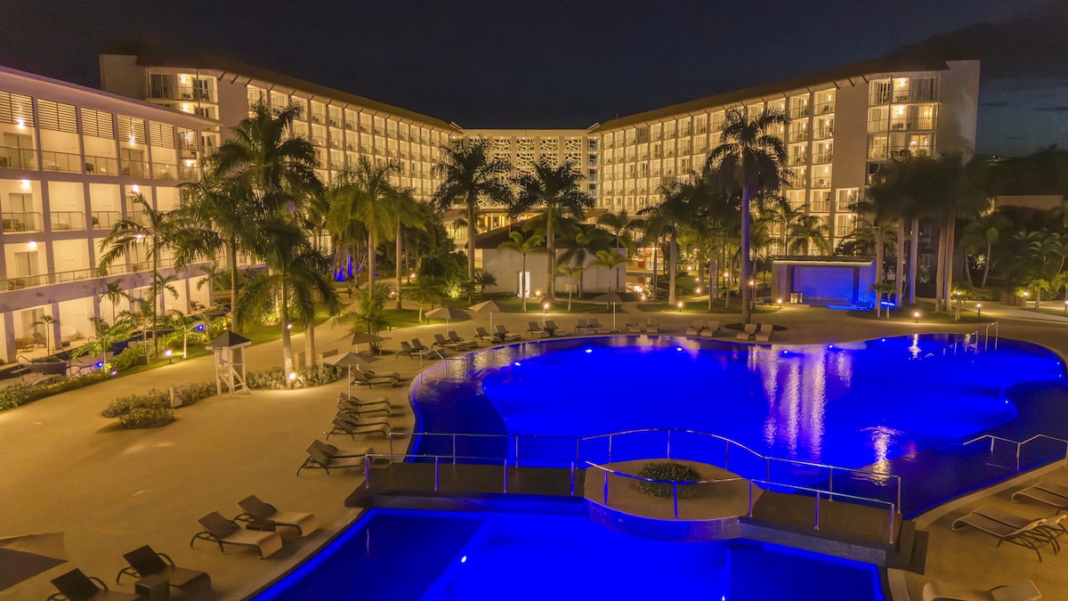 A hotel with a large, illuminated swimming pool surrounded by palm trees and lounge chairs.
