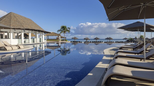 A pool area with lounge chairs and umbrellas, a building with a thatched roof, and a clear view of the ocean.