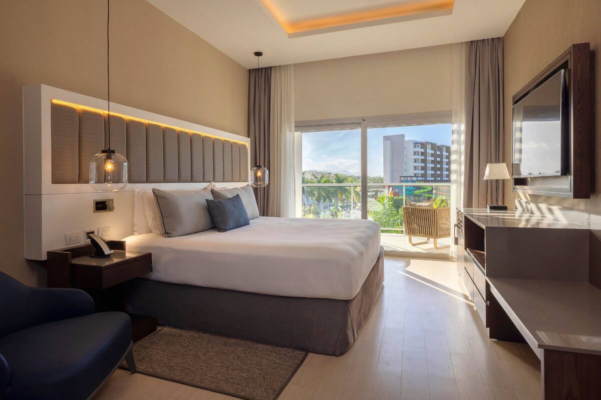 A modern hotel room with a large bed, a desk, a chair, a TV, and a balcony with a view of buildings and greenery.