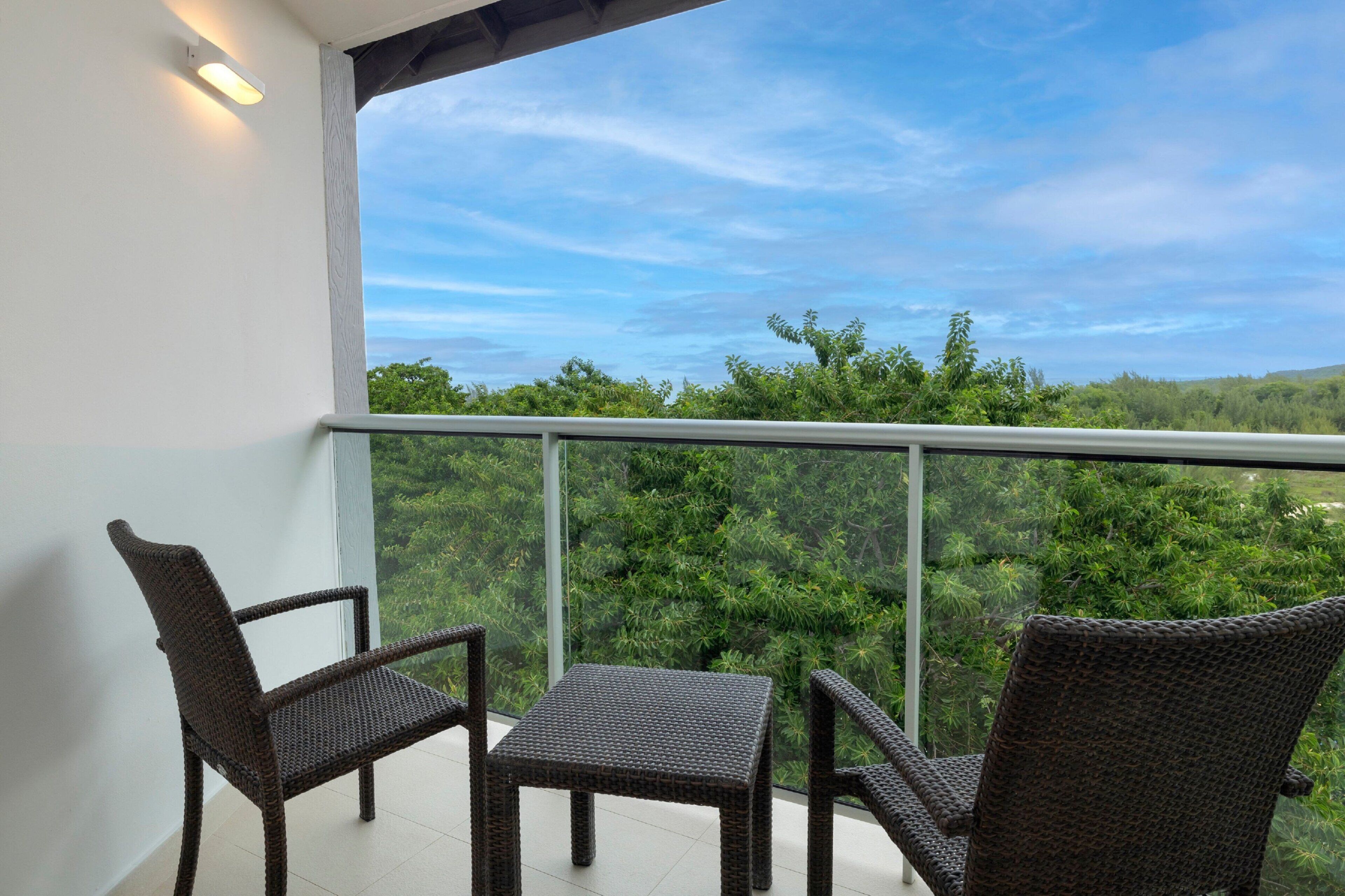 A balcony with wicker furniture and a glass railing, overlooking a lush green landscape.
