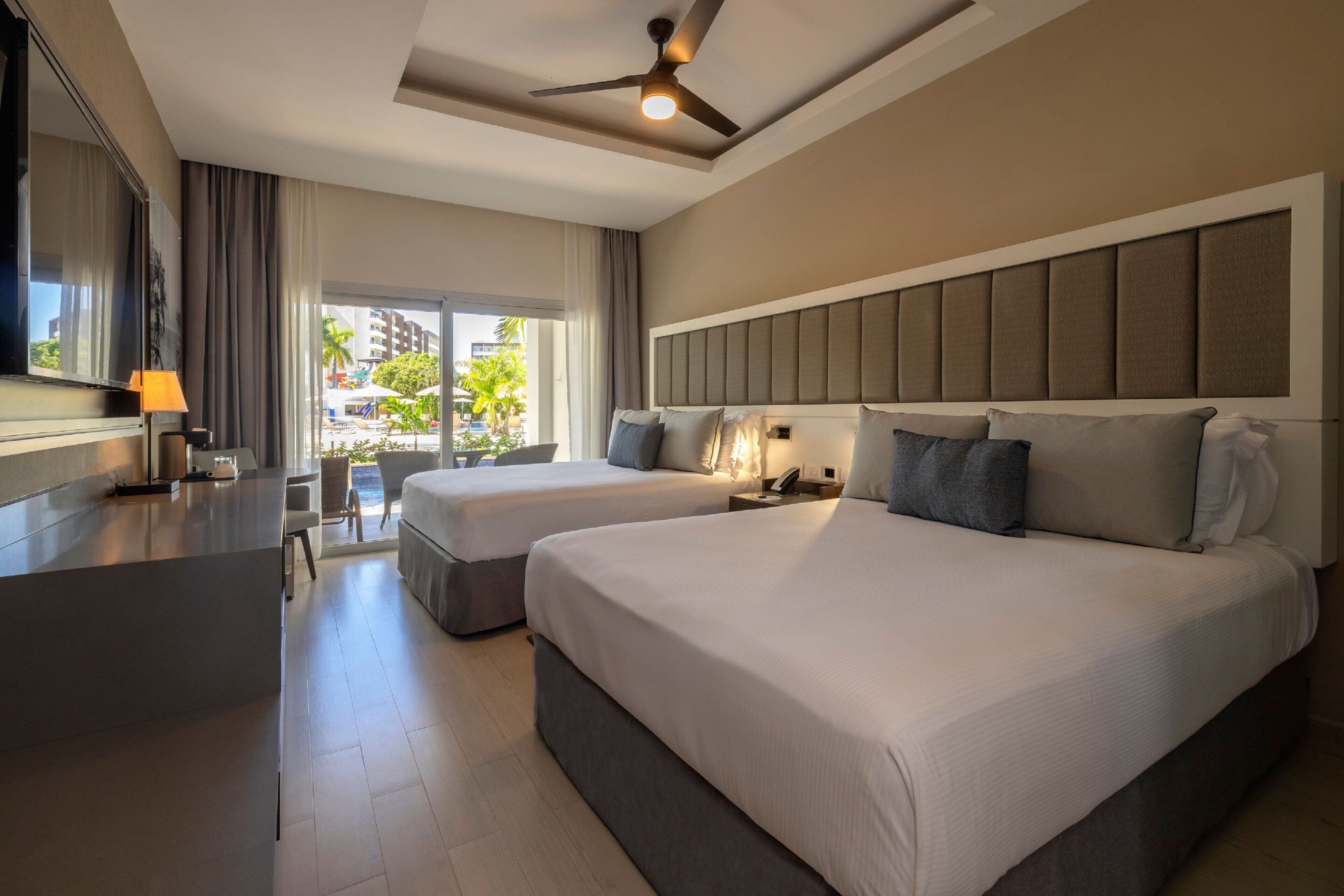 A hotel room with two beds, a desk, a ceiling fan, and a view of the outdoors.