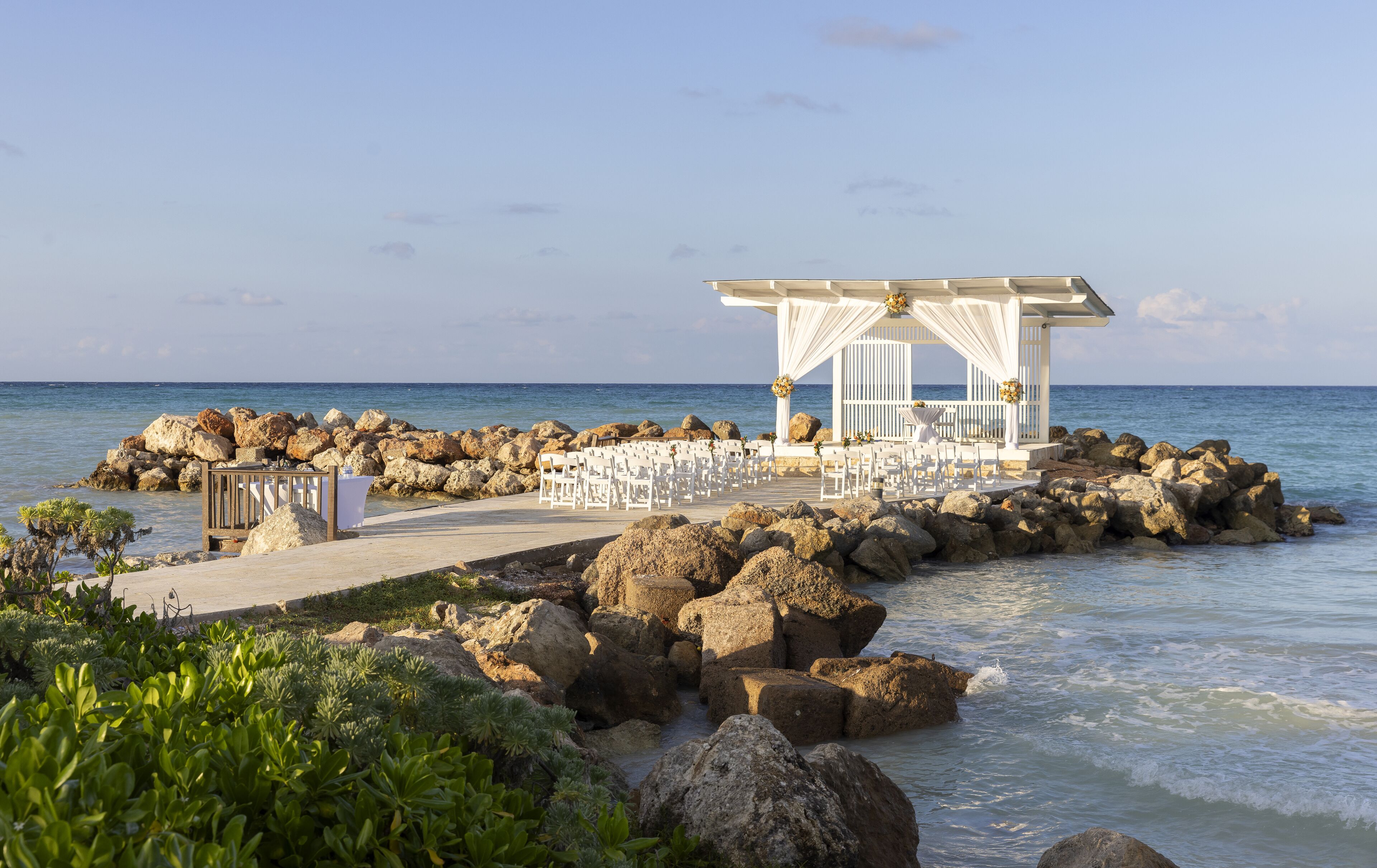 A beachside wedding venue with a white gazebo, rows of white chairs, and a rocky shoreline.