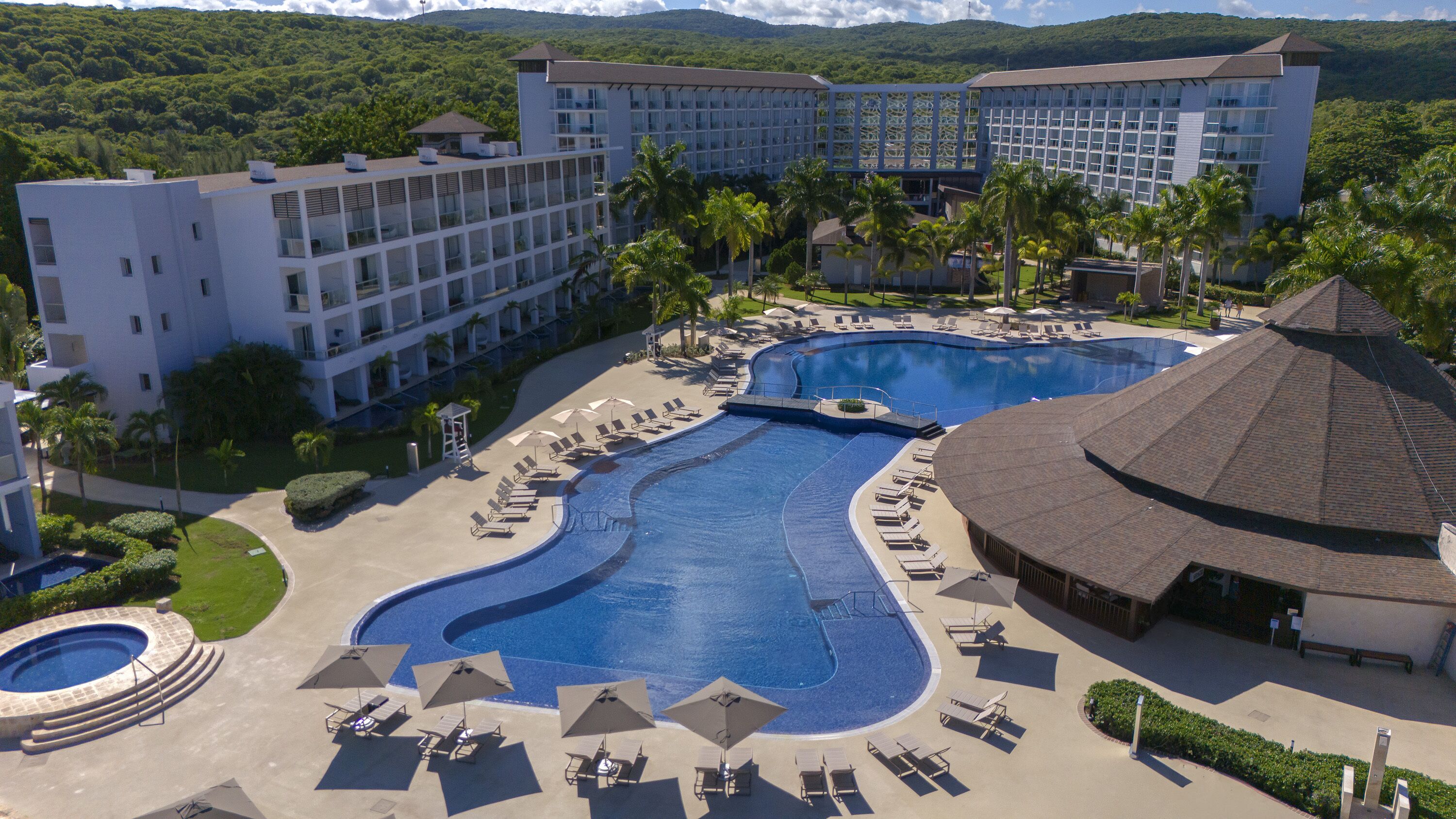 A resort with a large swimming pool, multiple lounge areas, and a view of the surrounding greenery.