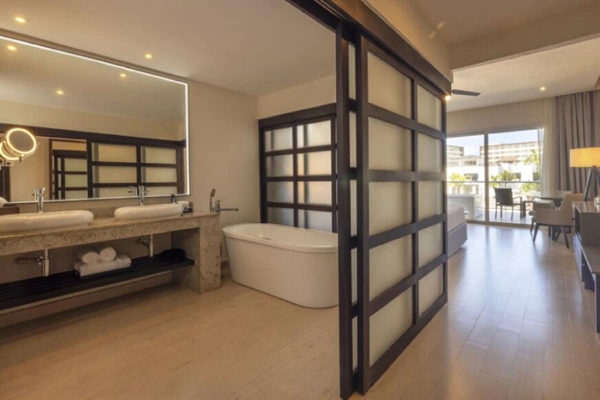 A modern bathroom with a large mirror, dual sinks, a bathtub, and a sliding glass door leading to a balcony.