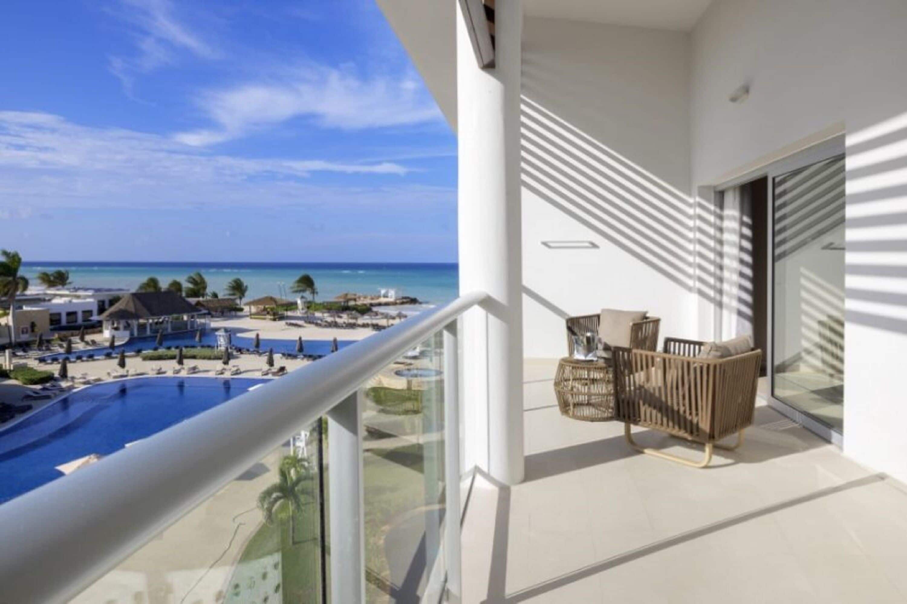 Un balcón con vista a la piscina, una zona de estar con mobiliario de mimbre y una puerta de cristal que da a la playa.