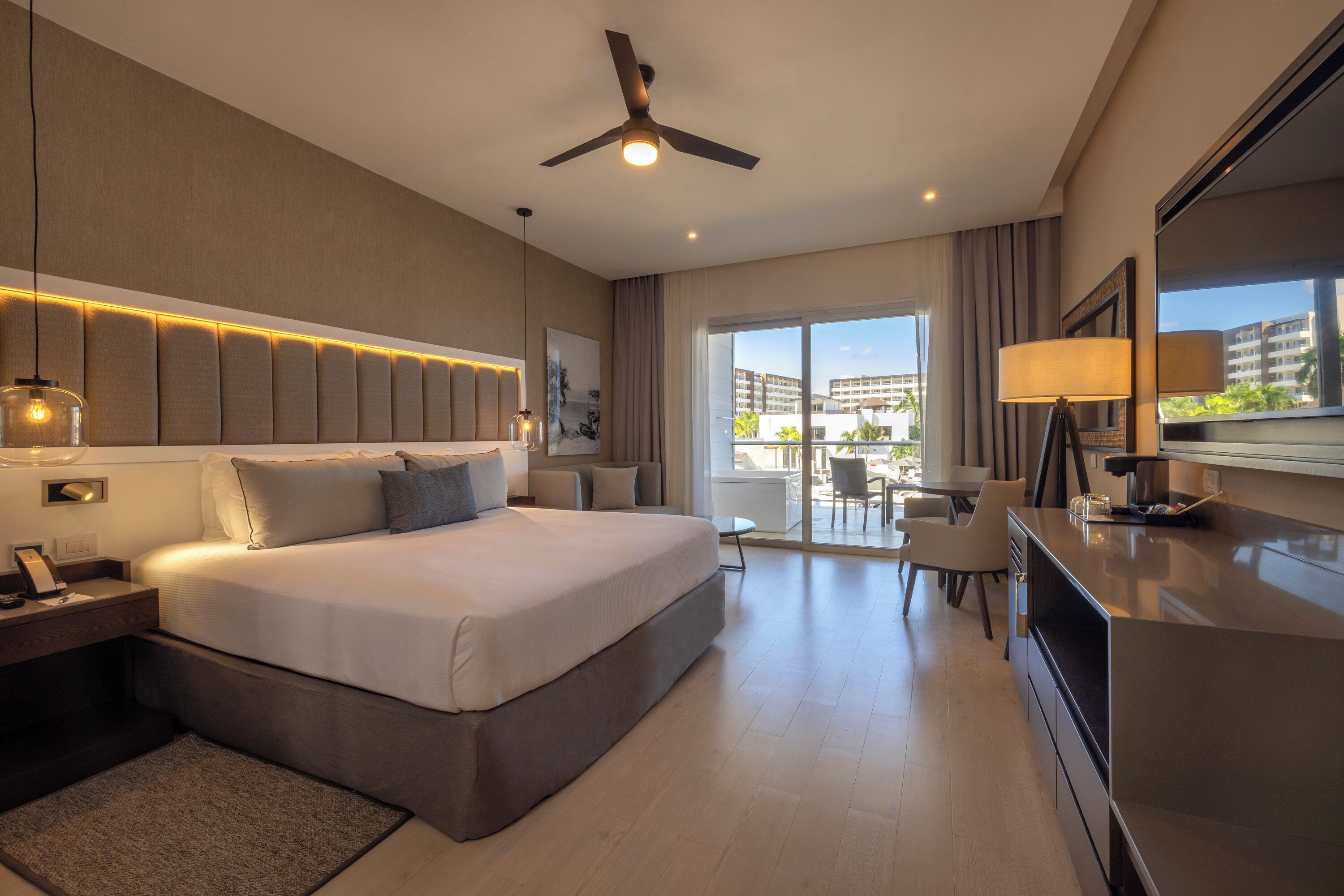 A modern hotel room with a large bed, a desk, and a balcony with a view of buildings.