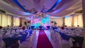 Une salle de banquet avec des rangées de tables recouvertes de nappes blanches et de chemins de table bleus, une allée bordée d’un tapis rouge et une scène dotée d’un éclairage coloré et de décorations florales.