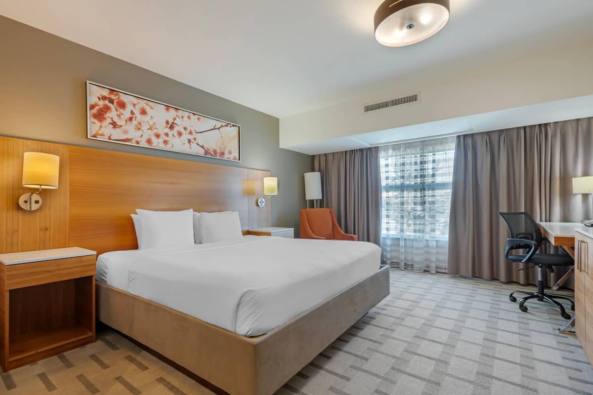 A hotel room with a large bed, a desk, a chair, a window with curtains, and a wall-mounted artwork.