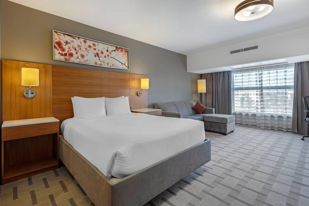 A hotel room with a large bed, two bedside tables with lamps, a sofa, and a large window with curtains.