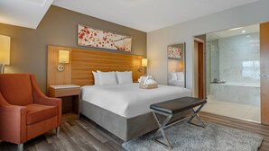 Habitación de hotel con cama, una silla, mesita de noche, lámpara, una obra de arte en la pared, baño con ducha de vidrio y pared de mármol.