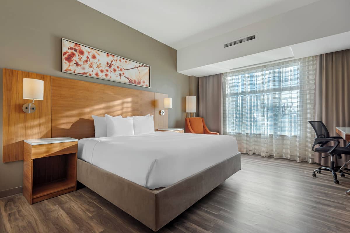 A modern hotel room with a large bed, two bedside tables with lamps, a wooden headboard, a large window with curtains, and a desk with a chair.