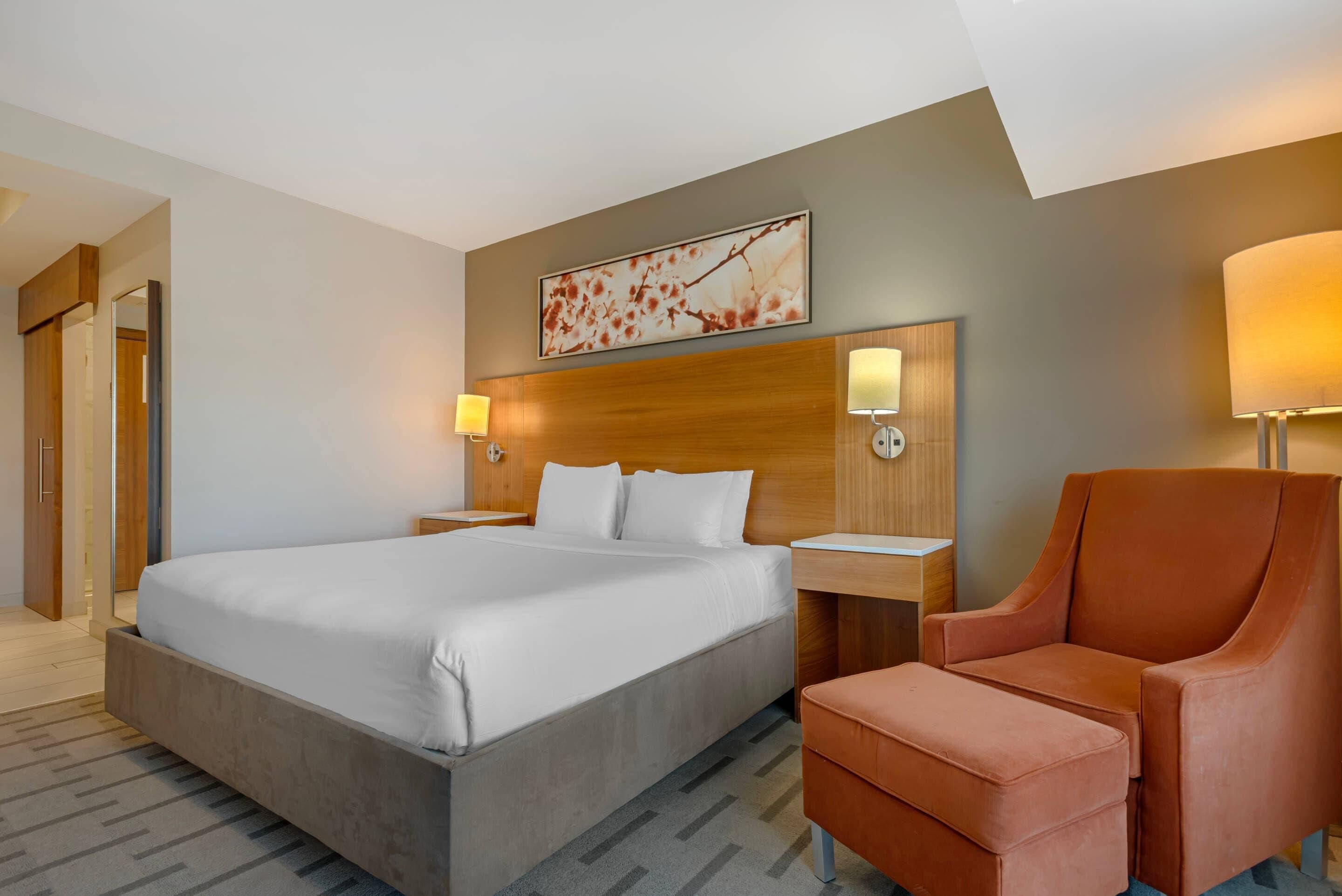 A hotel room with a large bed, a wooden headboard, two bedside lamps, a chair, and a wall-mounted artwork.