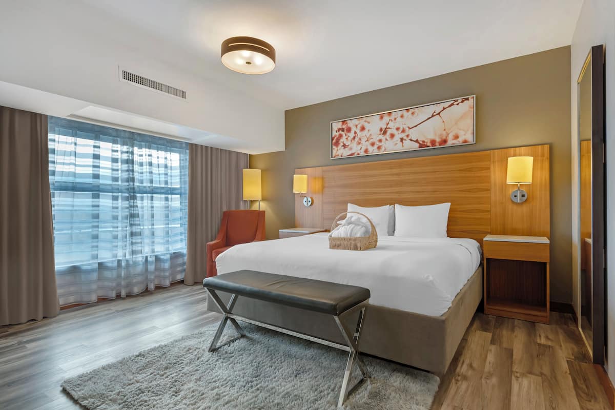 A hotel room with a large bed, a bench, two bedside lamps, a wall-mounted artwork, and a view through sheer curtains.