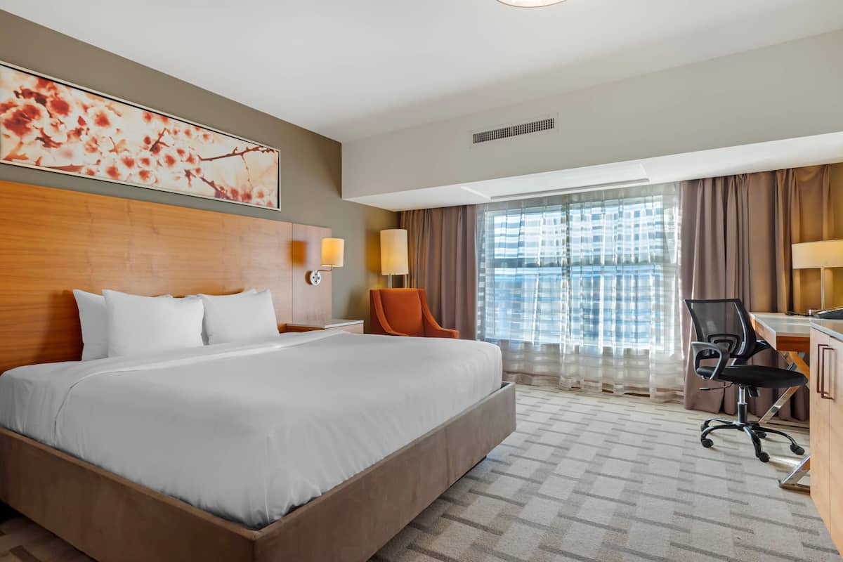 A hotel room with a large bed, a desk with a chair, a window with curtains, and a wall-mounted artwork.