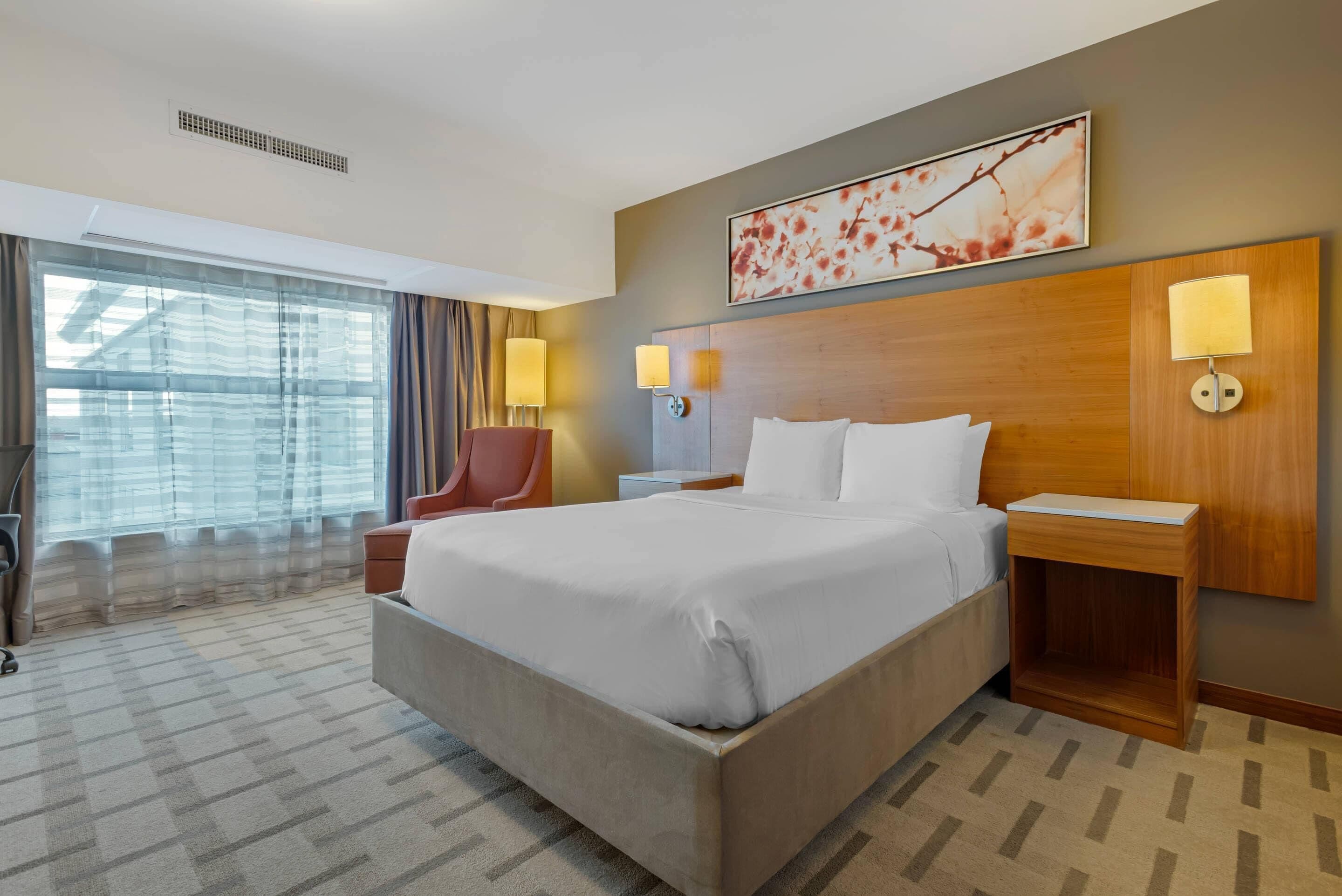 A hotel room with a large bed, two bedside lamps, a headboard, a window with curtains, and a wall-mounted artwork.
