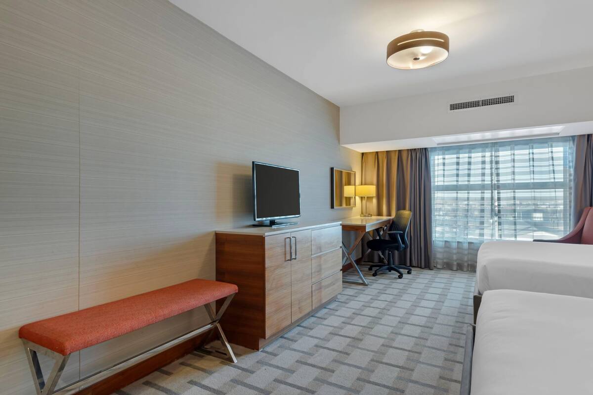 A hotel room with a bed, a desk with a chair, a television, a bench, and a window with curtains.