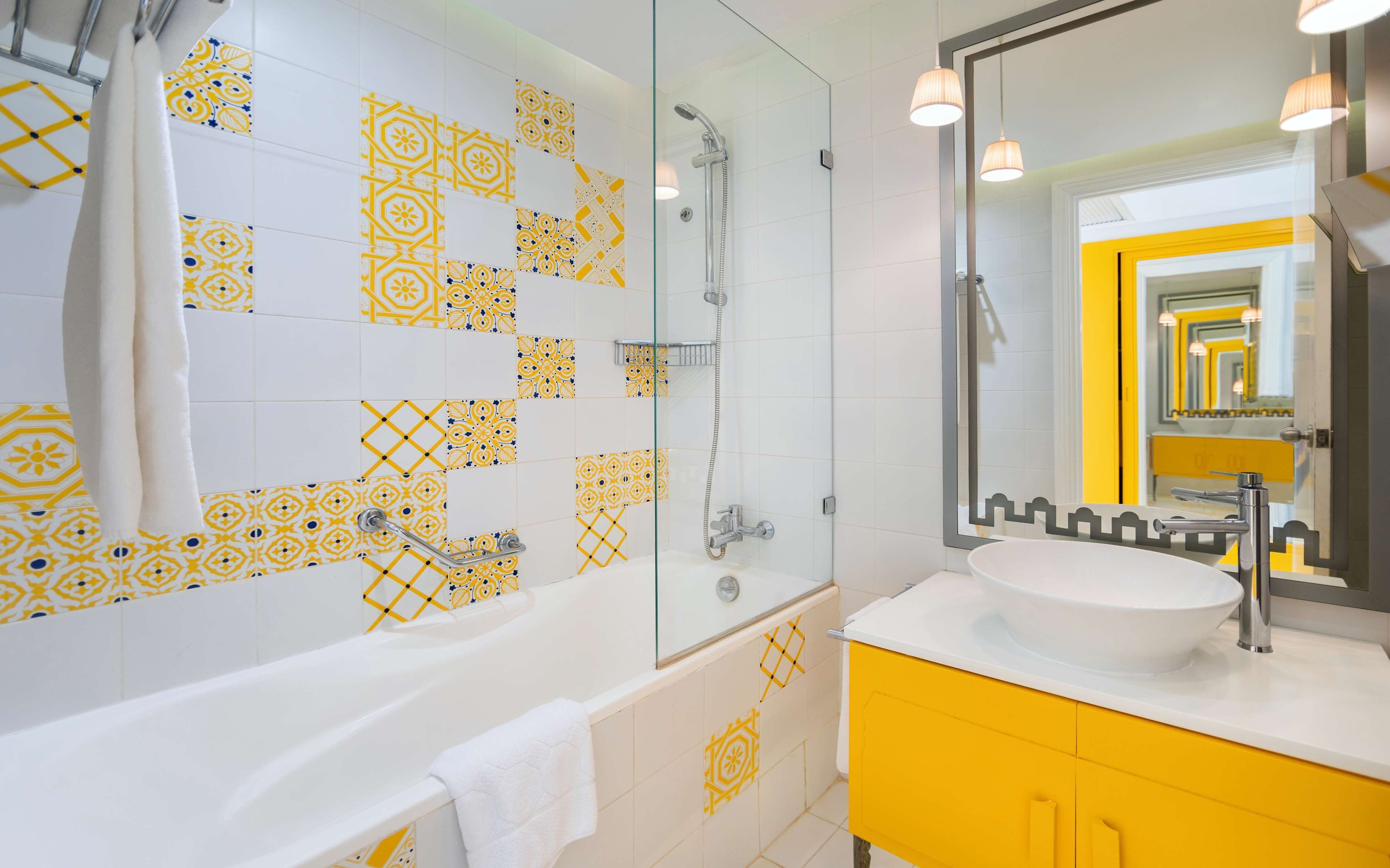 Une salle de bain moderne avec des carreaux à motifs, une douche en verre, une baignoire blanche, un meuble-lavabo jaune et un grand miroir.