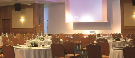 Un salón de conferencias con mesas redondas, sillas y una pantalla grande para presentaciones.