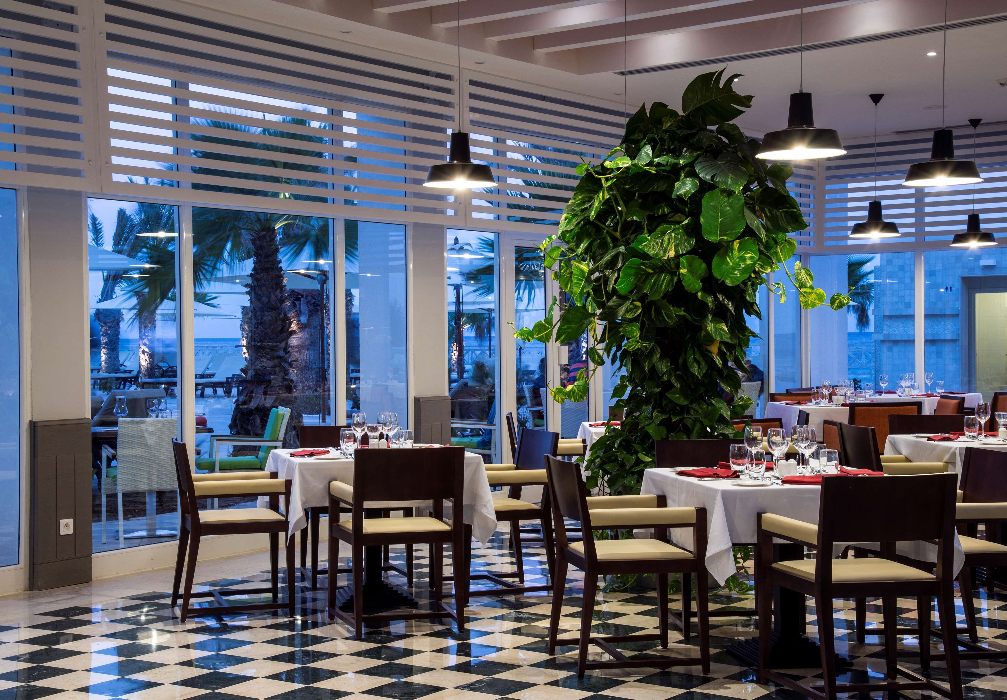 A restaurant with checkered flooring, white tables with red napkins, and large windows overlooking palm trees.
