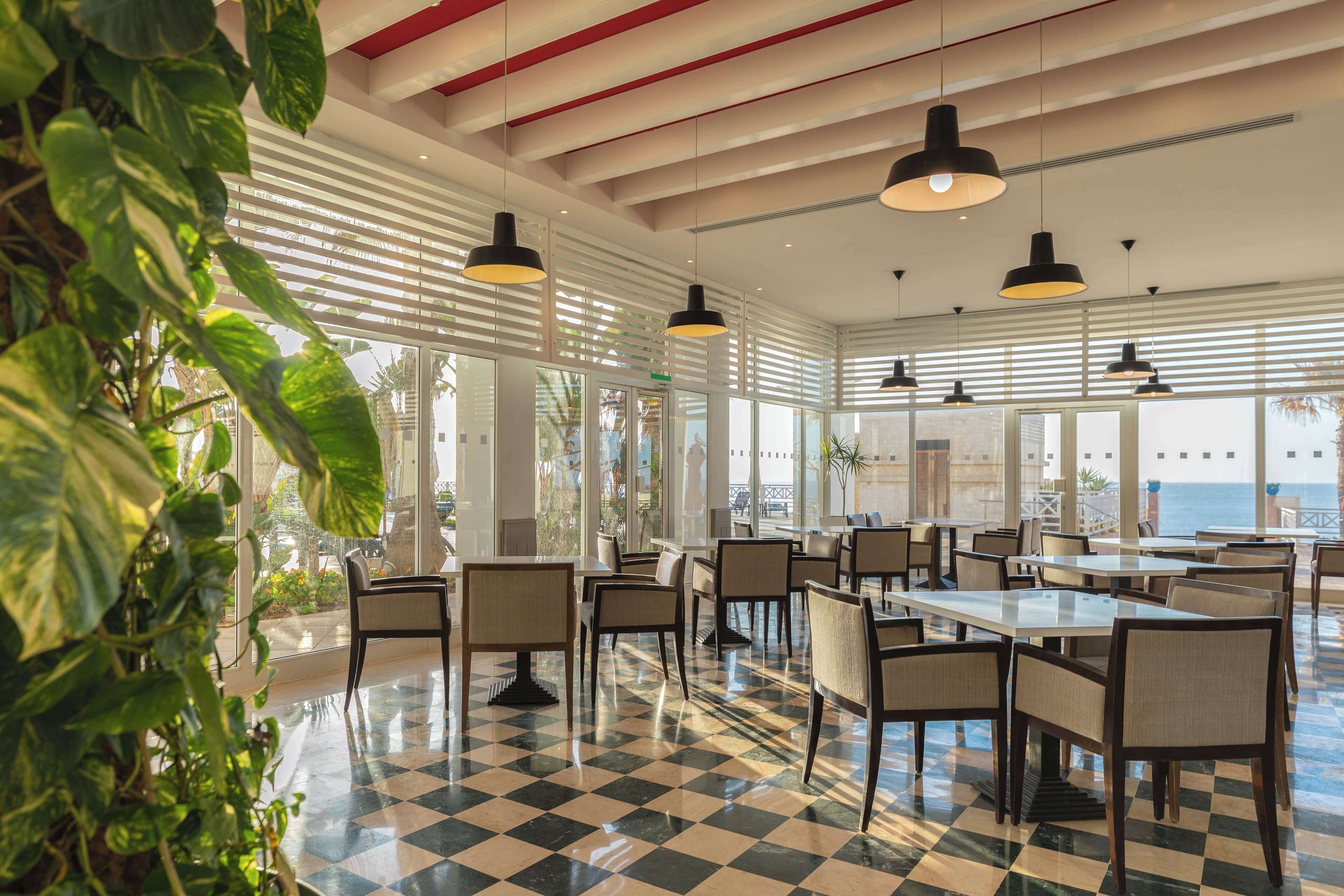 A spacious indoor dining area with checkered flooring, multiple tables and chairs, hanging pendant lights, and large windows offering views of the ocean.